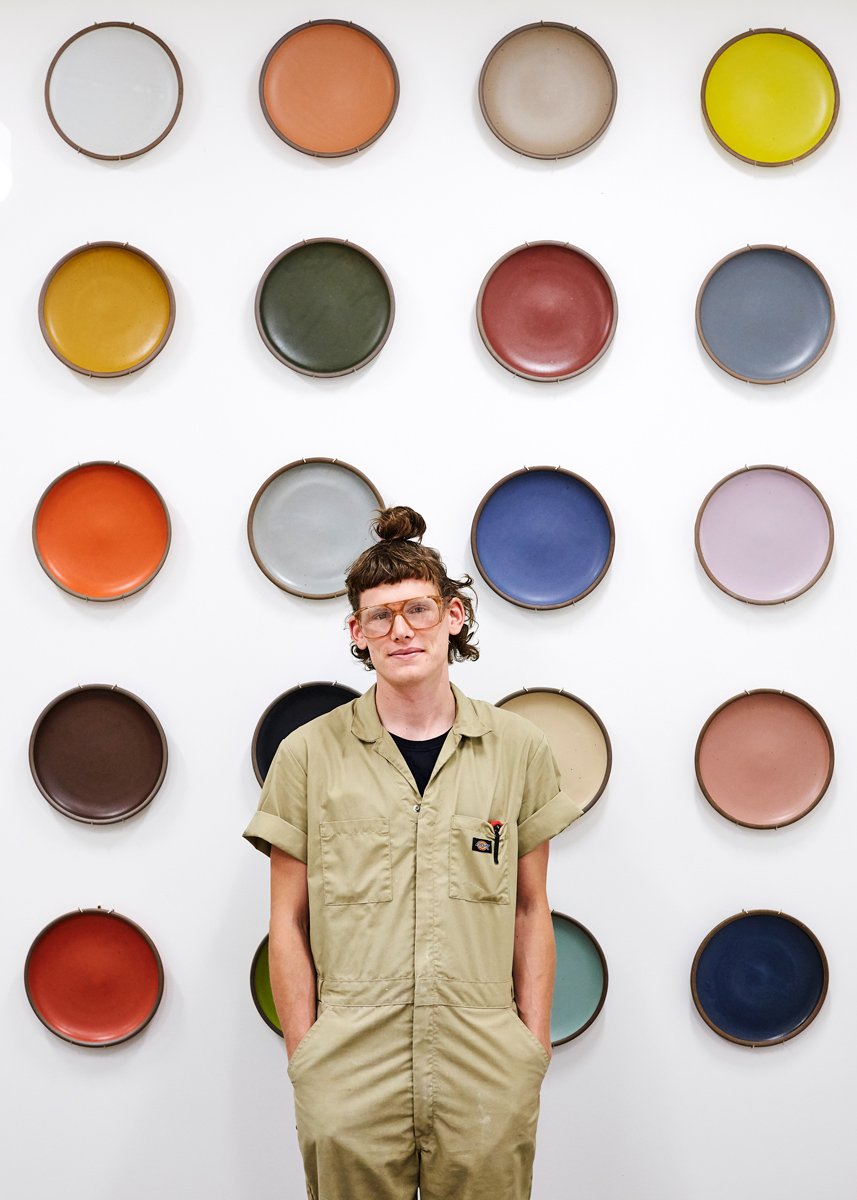Kyle Crowder, wearing a tan jumpsuit and safety glasses, poses in front of a wall with dinner plates hanging in many of East Fork Pottery's historical glaze colorways